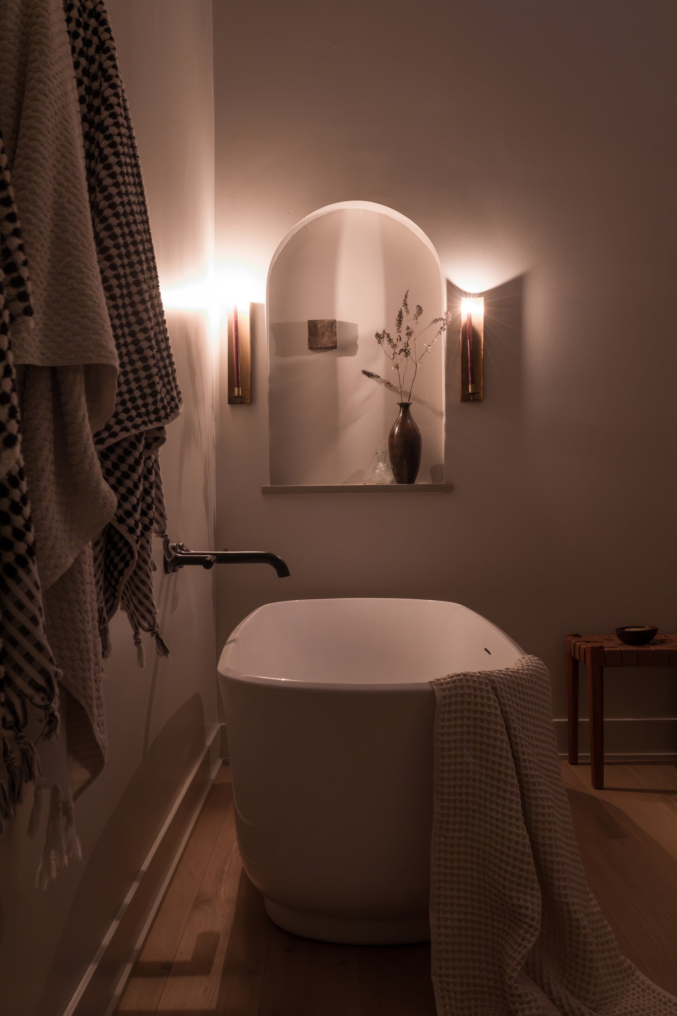 Arched bathroom niche styled with a ceramic vase, dried florals, and brass sconces
