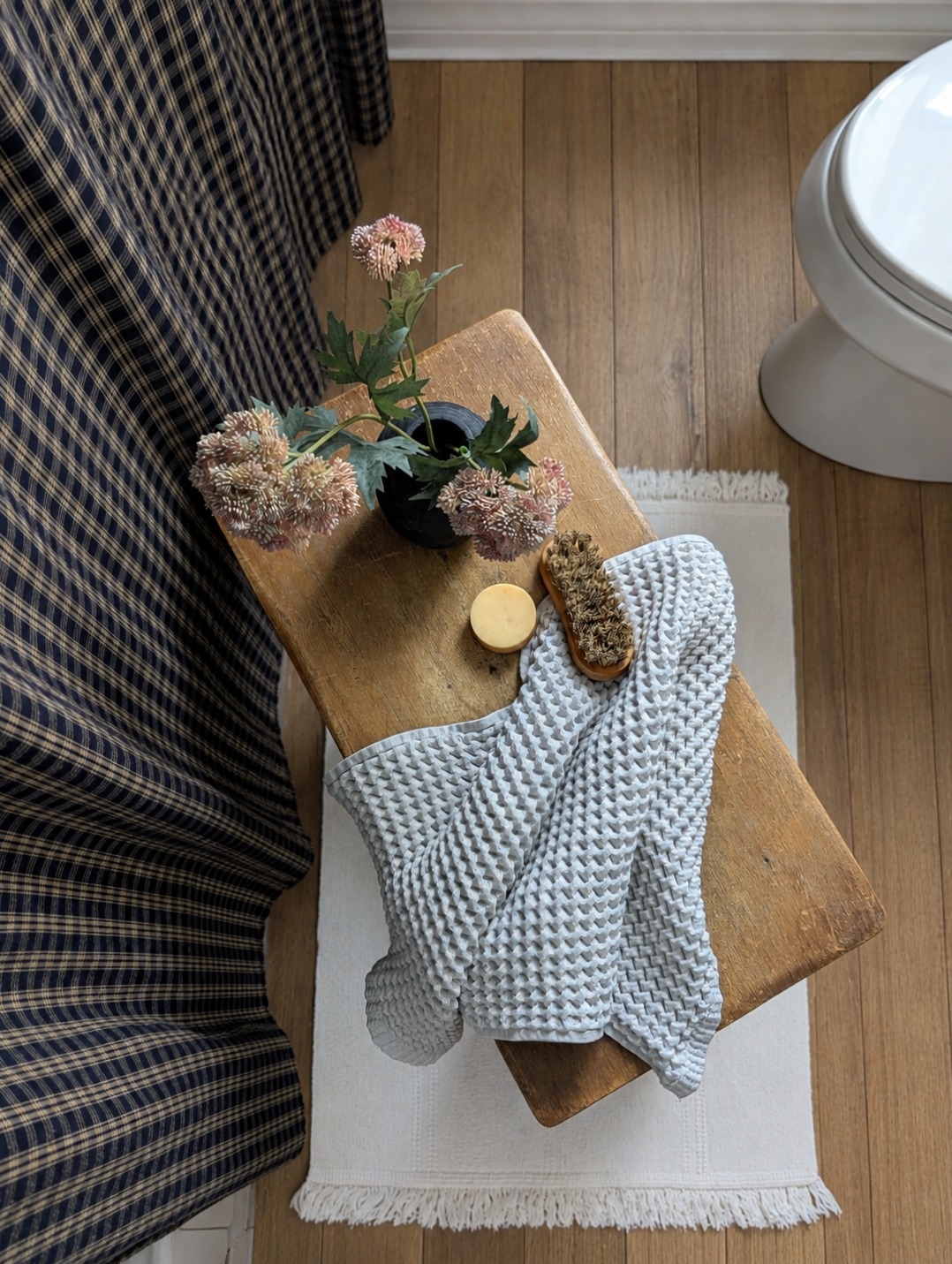 Overhead flatlay of a wooden stool styled with a waffle towel, dried flowers, and bath brush