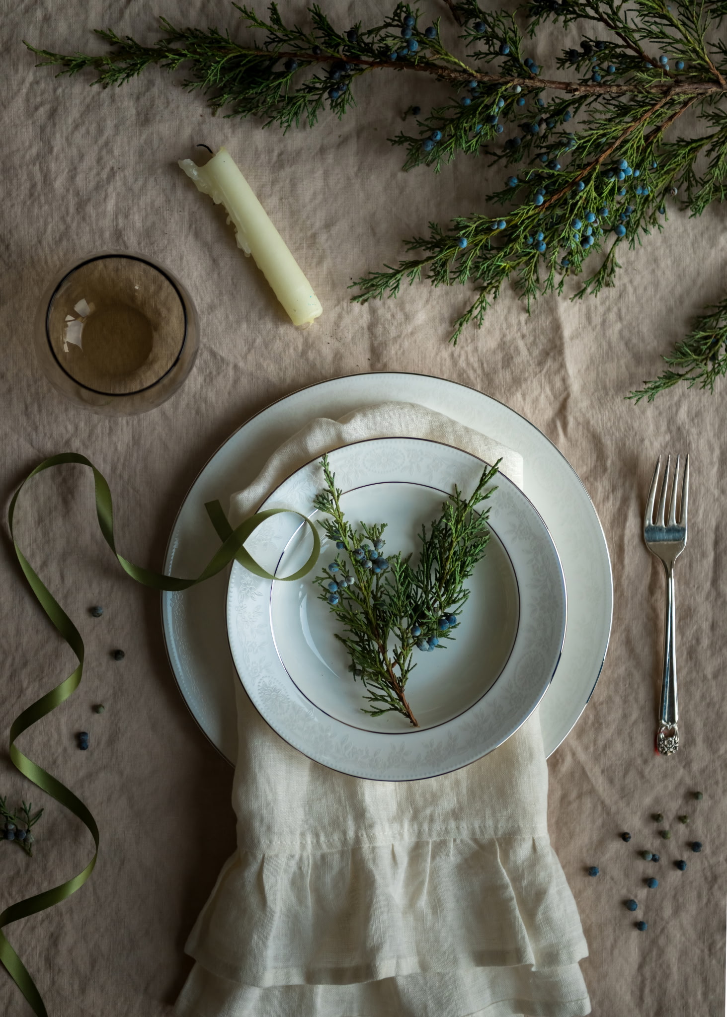 Overhead view of a styled table setting with silver-rimmed plates, juniper sprigs, and ribbon